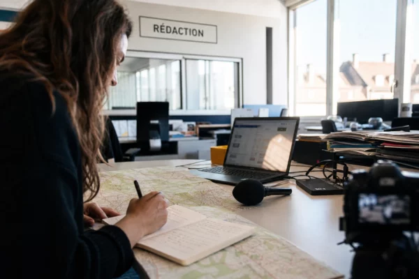 Un journaliste prépare un sujet climat dans une rédaction lumineuse avec carnet, carte floue et micro sans logo.