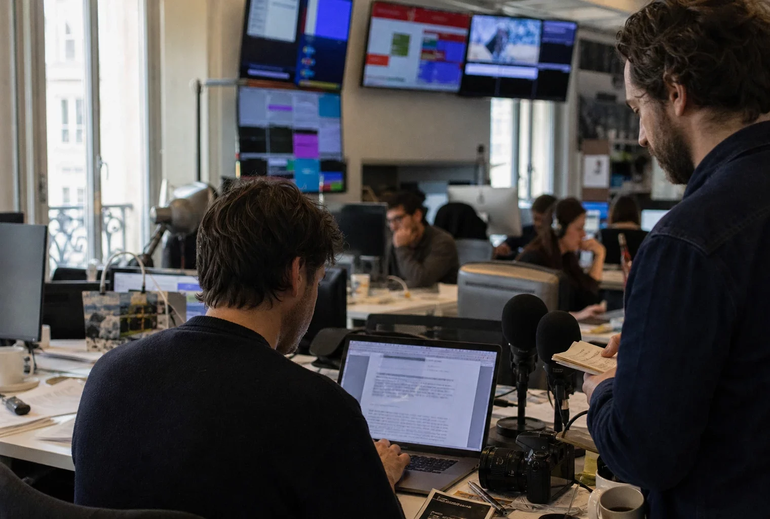 Dans une rédaction parisienne, un journaliste et un éditeur préparent un sujet diplomatique devant écrans flous et micros.