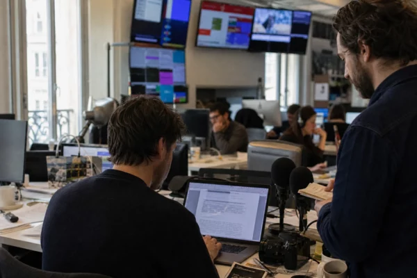 Dans une rédaction parisienne, un journaliste et un éditeur préparent un sujet diplomatique devant écrans flous et micros.