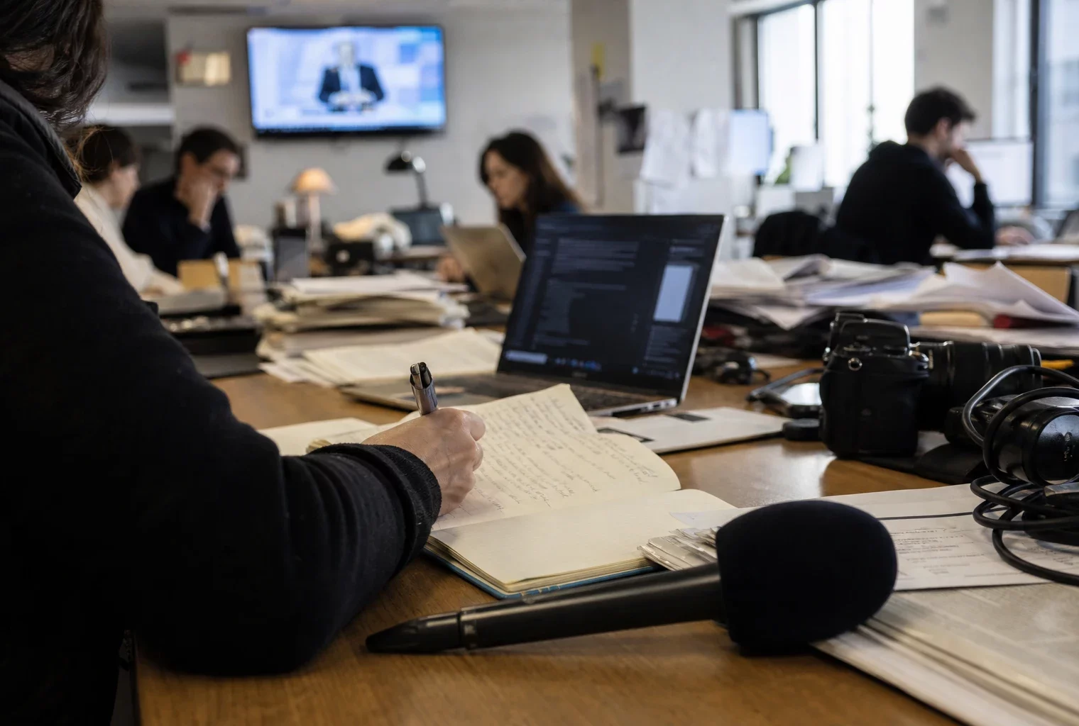Rédaction française en pleine enquête politique, journaliste prenant des notes près d’un micro et d’écrans flous.