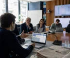 Réunion de rédaction dans une newsroom française lumineuse avec journaliste, écrans flous et matériel de presse.