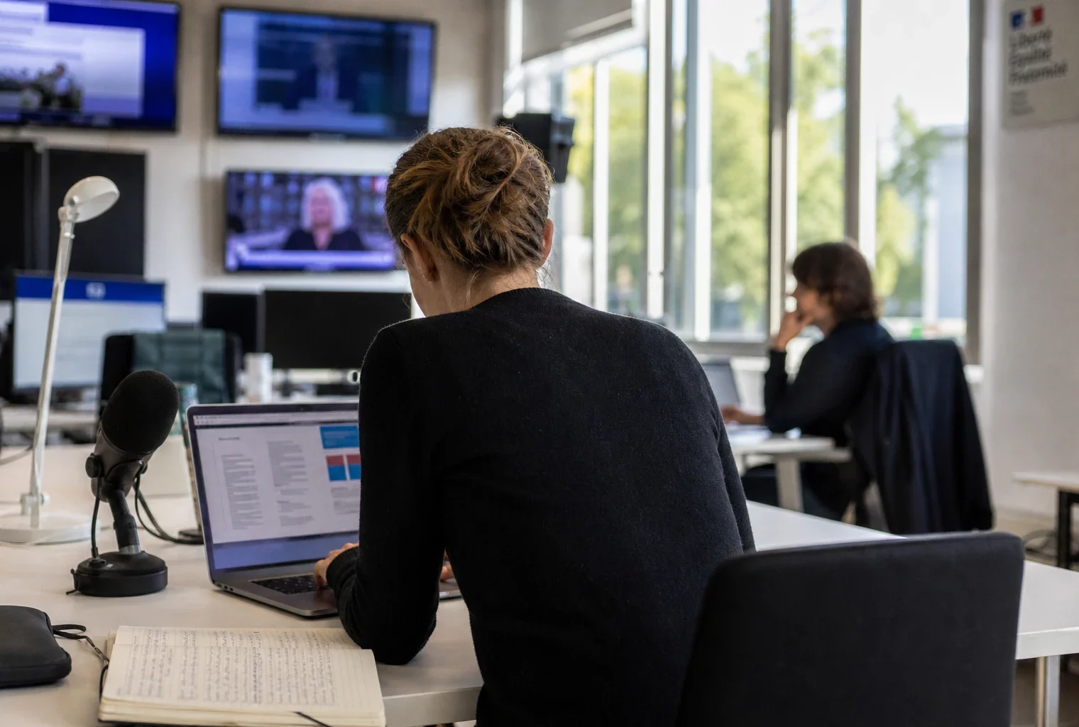 Journaliste dans une rédaction française préparant un sujet sur les intercommunalités, avec micro, carnet et écrans flous.