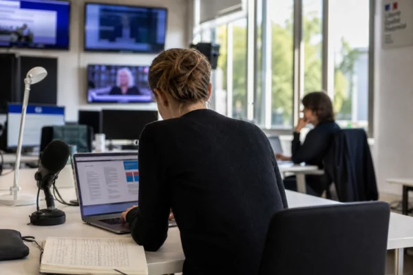 Journaliste dans une rédaction française préparant un sujet sur les intercommunalités, avec micro, carnet et écrans flous.