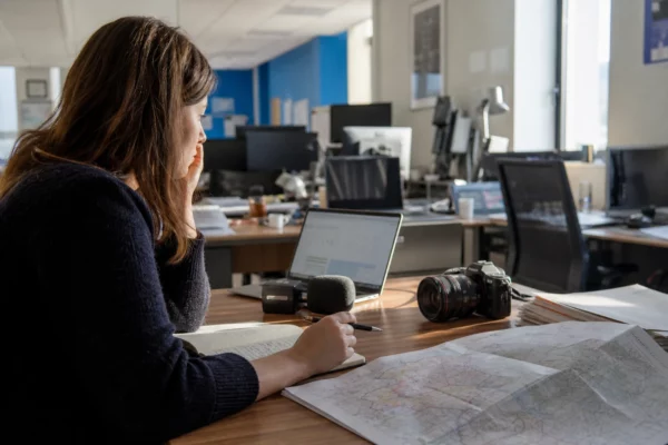 Journaliste en rédaction préparant un sujet territorial avec carnet, micro sans logo et carte papier floue.