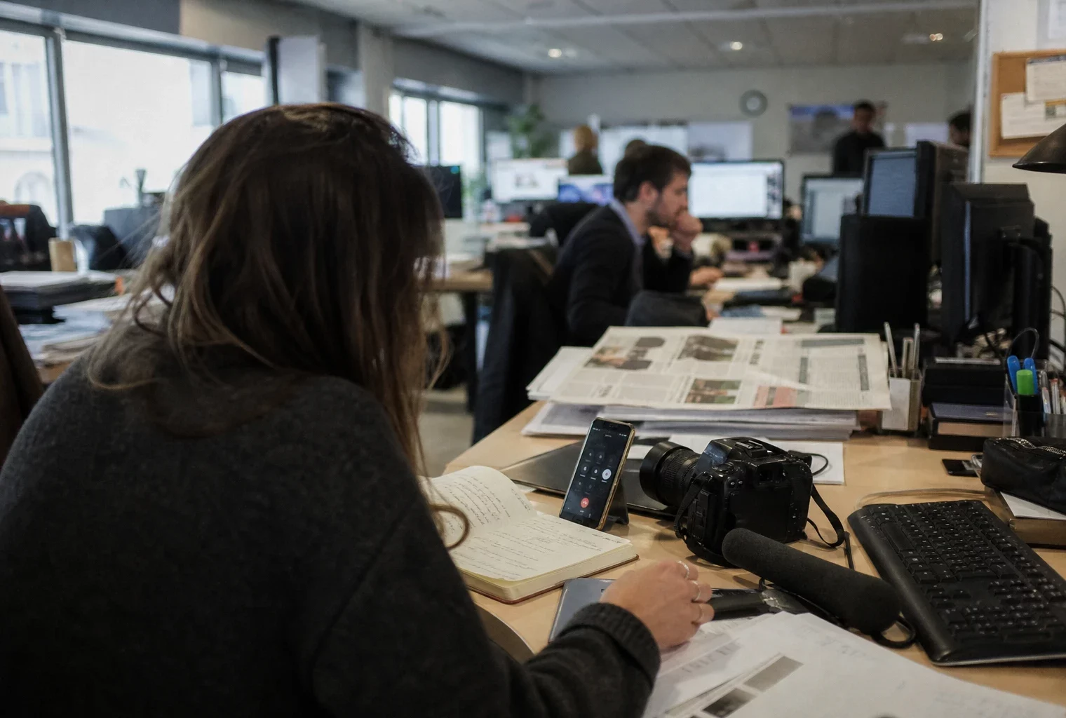 Journaliste dans une rédaction parisienne, avec micro, caméra et écrans flous dans une ambiance claire et contemporaine.