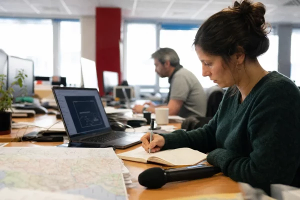 Journaliste en rédaction locale préparant un sujet politique territorial avec carnet, micro sans logo et écran flou.