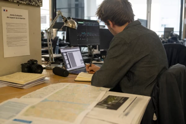 Journaliste en rédaction française préparant un sujet politique, avec carnet, micro sans logo et documents flous sur la table.