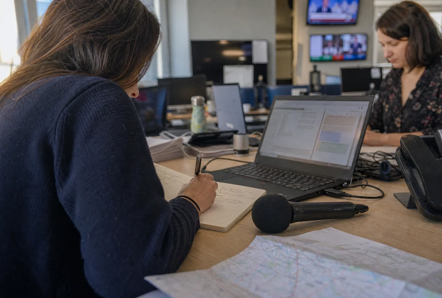 Journaliste en rédaction préparant un sujet territorial avec carnet, micro et carte papier floue.