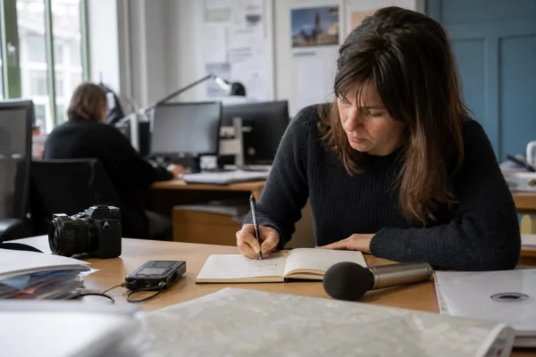 Journaliste en rédaction préparant un sujet local avec carnet, micro sans logo et carte papier floue.