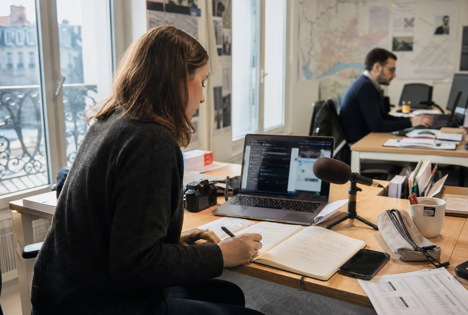 Journaliste local préparant un sujet territorial dans une rédaction française, avec carnet, micro et écran flou.