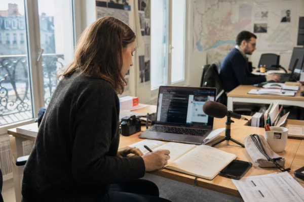 Journaliste local préparant un sujet territorial dans une rédaction française, avec carnet, micro et écran flou.