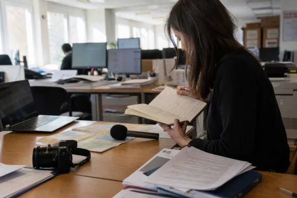 Journaliste en rédaction locale préparant un sujet parlementaire, carnet ouvert, micro sans logo et écrans flous.