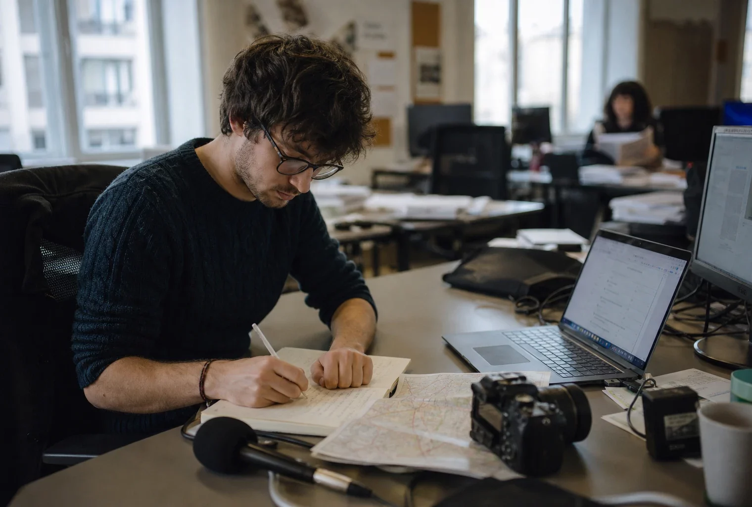 Journaliste en rédaction préparant un sujet territorial, avec carnet, micro sans logo et écran flou en arrière-plan.