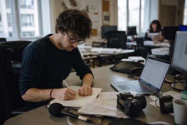 Journaliste en rédaction préparant un sujet territorial, avec carnet, micro sans logo et écran flou en arrière-plan.
