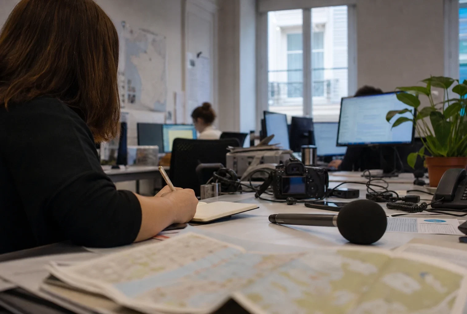 Journaliste dans une rédaction locale préparant un sujet politique, carnet ouvert, micro sans logo et cartes floues.