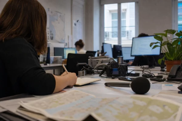 Journaliste dans une rédaction locale préparant un sujet politique, carnet ouvert, micro sans logo et cartes floues.