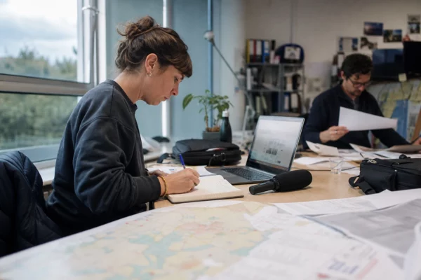 Journaliste en rédaction locale préparant un sujet politique avec carnet, micro et carte papier floue