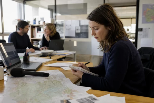 Journaliste dans une rédaction locale préparant un sujet territorial avec micro, carnet et carte floue.