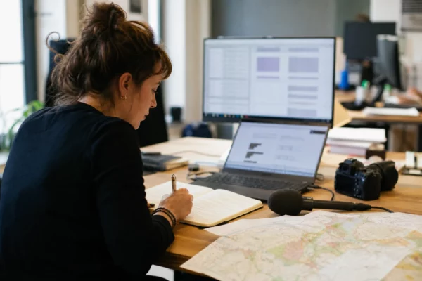 Journaliste en rédaction locale préparant un sujet territorial avec carnet, carte floue et micro sans logo.