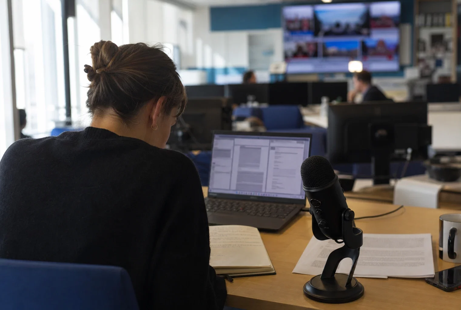 Journaliste dans une rédaction lumineuse avec carnet, micro et écran abstrait préparant un sujet politique.