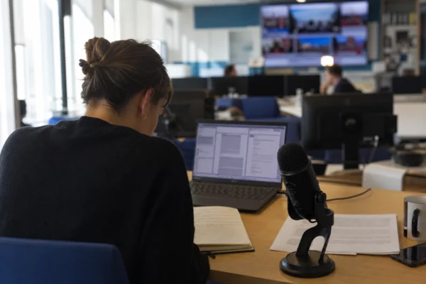 Journaliste dans une rédaction lumineuse avec carnet, micro et écran abstrait préparant un sujet politique.