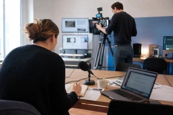 Dans une rédaction française, des journalistes préparent un sujet politique avec micros, carnet et écrans flous.
