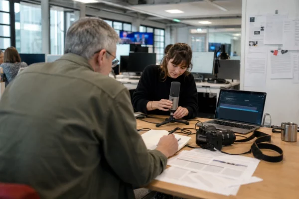 Rédaction politique française préparant un sujet sur la présidentielle 2027, avec micro, carnet et écran flou.