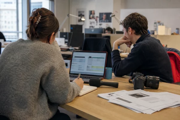 Deux journalistes en rédaction préparent un sujet sur la santé mentale des jeunes, avec micro, ordinateur et notes floues.