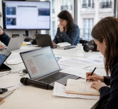 Rédaction française préparant un sujet politique, avec micro, carnet et écrans flous sans texte lisible.
