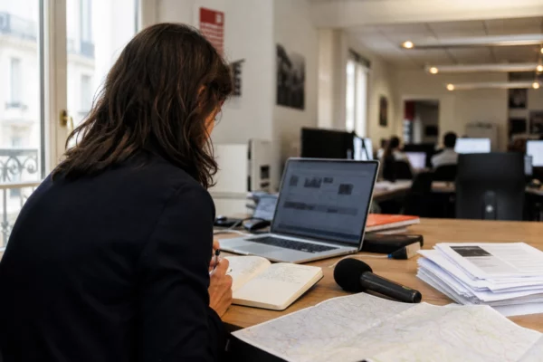 Journaliste en rédaction préparant un sujet politique, carnet ouvert, micro sans logo et carte floue sur la table.