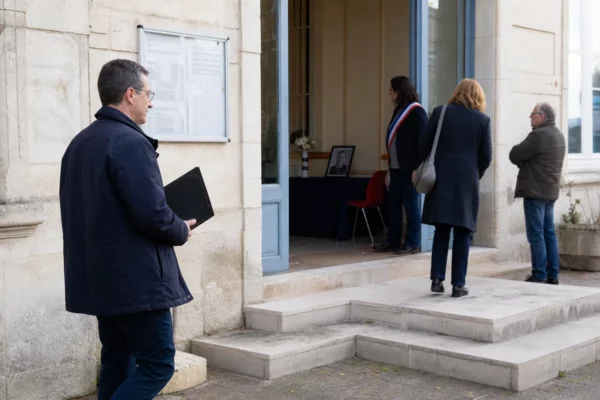 Des anonymes déposent un mot dans un registre de condoléances devant une mairie française, sous une lumière claire.
