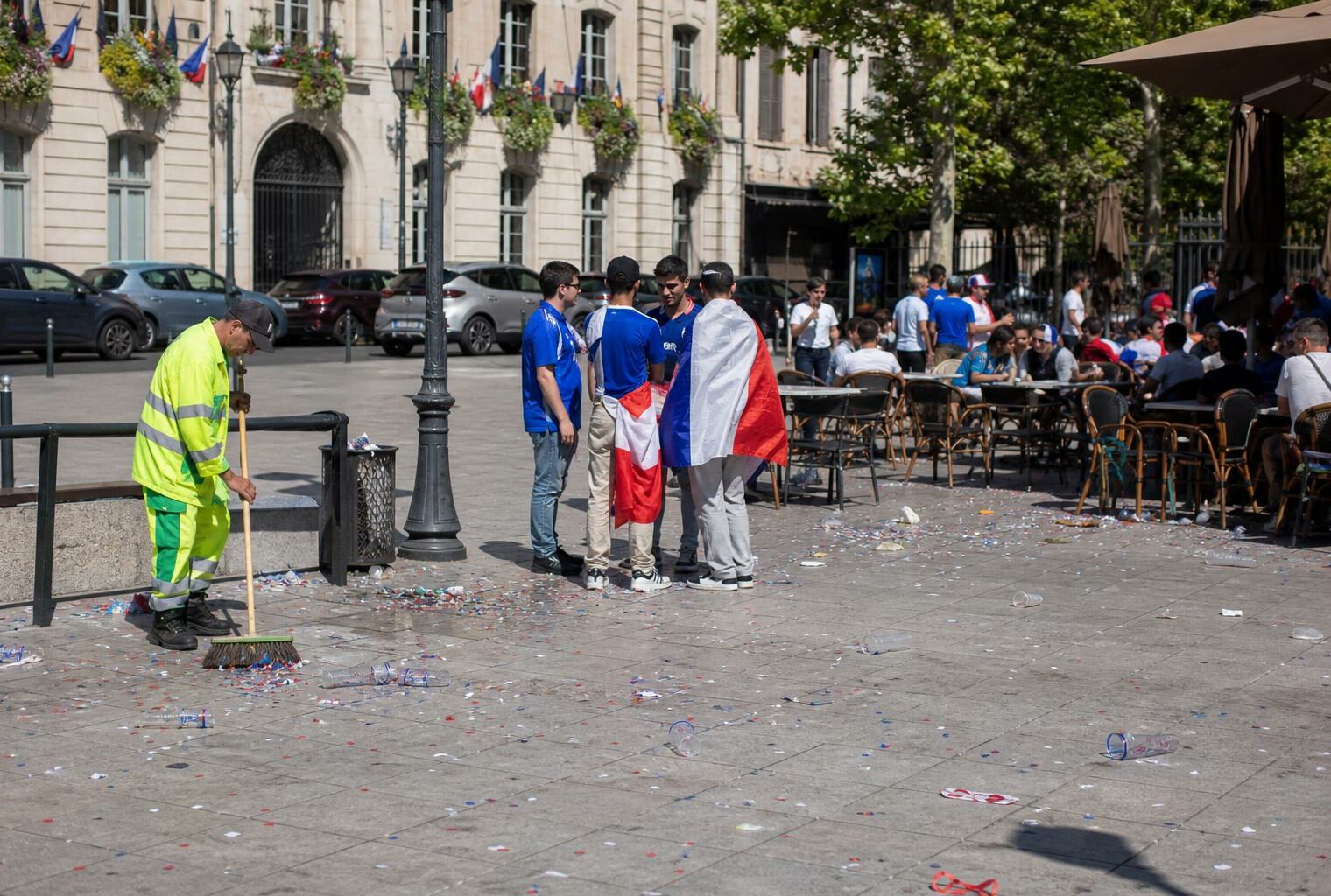 Place de ville française avec habitants anonymes et agent municipal nettoyant les traces d’une célébration
