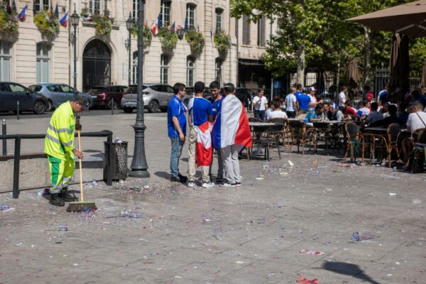 Place de ville française avec habitants anonymes et agent municipal nettoyant les traces d’une célébration