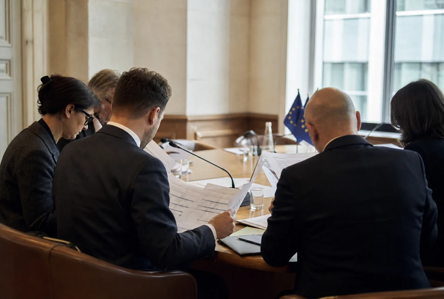 Réunion européenne sobre avec quatre délégués anonymes, dossiers sur table et drapeaux de table discrets.