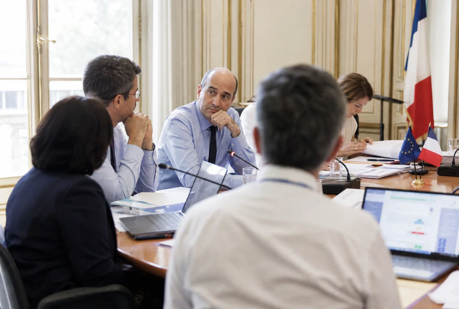 Réunion européenne sobre avec silhouettes de dos, dossiers ouverts et drapeaux de table discrets dans une salle claire.