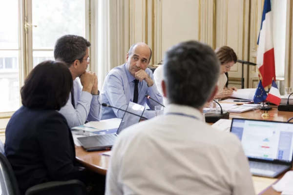 Réunion européenne sobre avec silhouettes de dos, dossiers ouverts et drapeaux de table discrets dans une salle claire.