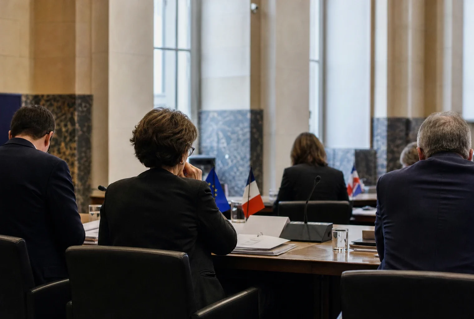 Réunion européenne sobre avec dossiers et petites drapeaux de table, vue de dos en salle institutionnelle claire.