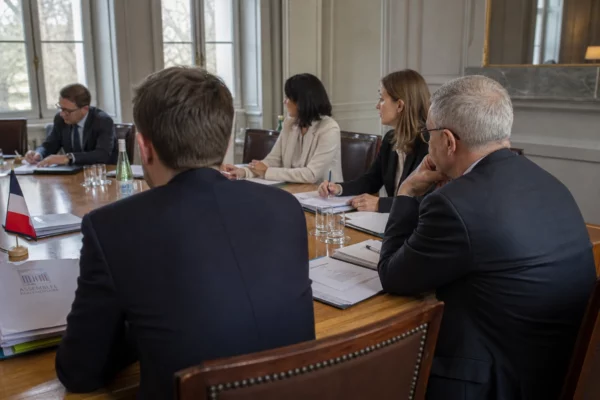 Réunion européenne sobre avec dossiers, silhouettes de dos et drapeaux discrets autour d’une table de négociation.