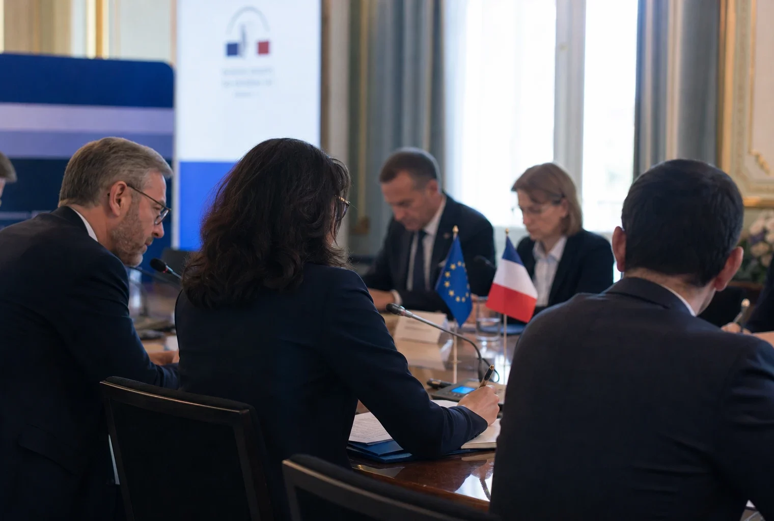 Réunion européenne sobre avec plusieurs participants, dossiers, micros et petits drapeaux de table dans une salle lumineuse.