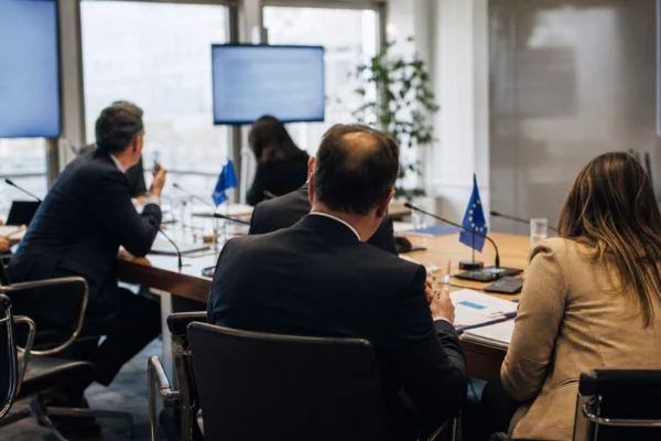 Réunion européenne sobre avec dossiers, micros et drapeaux de table discrets dans une salle institutionnelle lumineuse.