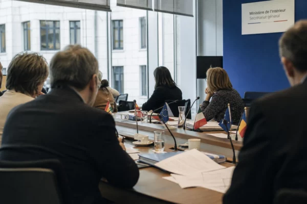 Réunion européenne sobre avec silhouettes de dos, dossiers et micros autour d’une table de négociation.