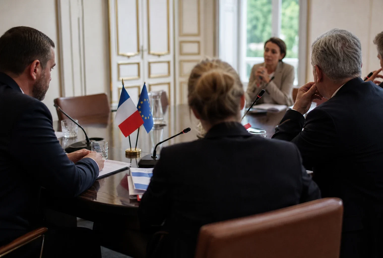 Réunion européenne sobre avec silhouettes anonymes, dossiers et micros, lumière claire et drapeaux de table discrets.
