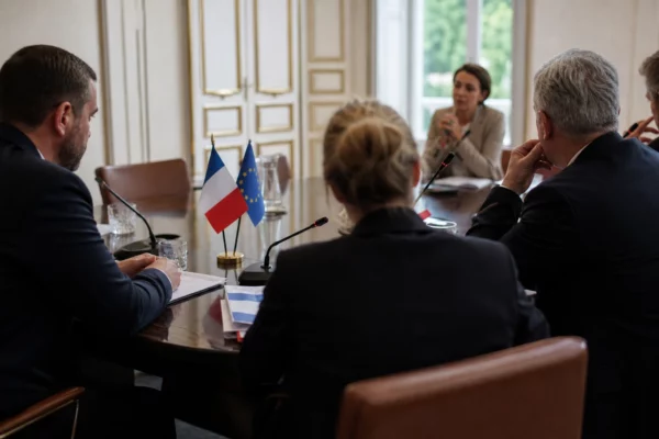 Réunion européenne sobre avec silhouettes anonymes, dossiers et micros, lumière claire et drapeaux de table discrets.