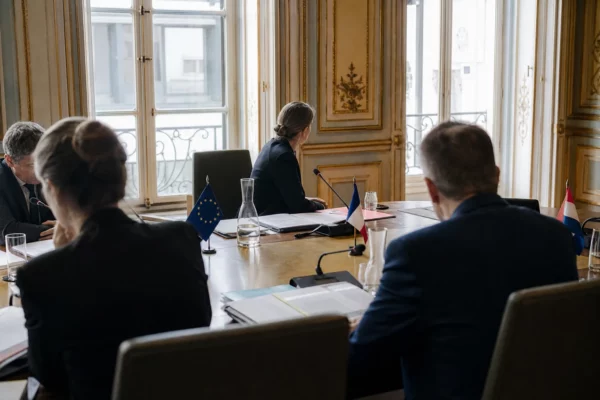 Réunion européenne sobre avec silhouettes anonymes, dossiers ouverts et micros sur une table de négociation.