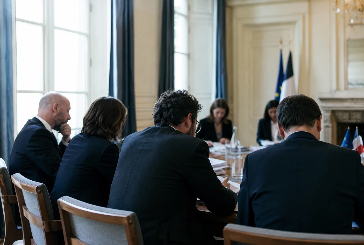 Réunion européenne sobre avec dossiers, silhouettes de dos et drapeaux de table discrets dans une lumière claire.