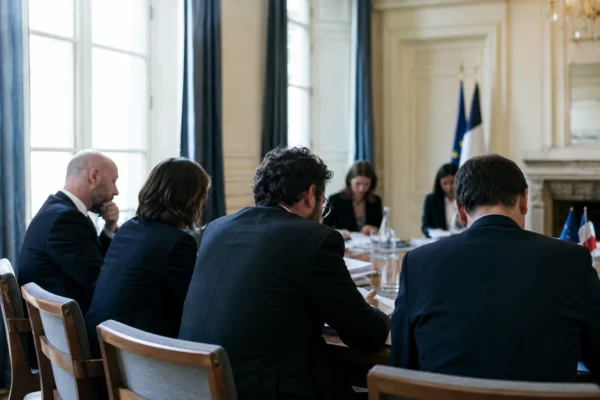 Réunion européenne sobre avec dossiers, silhouettes de dos et drapeaux de table discrets dans une lumière claire.