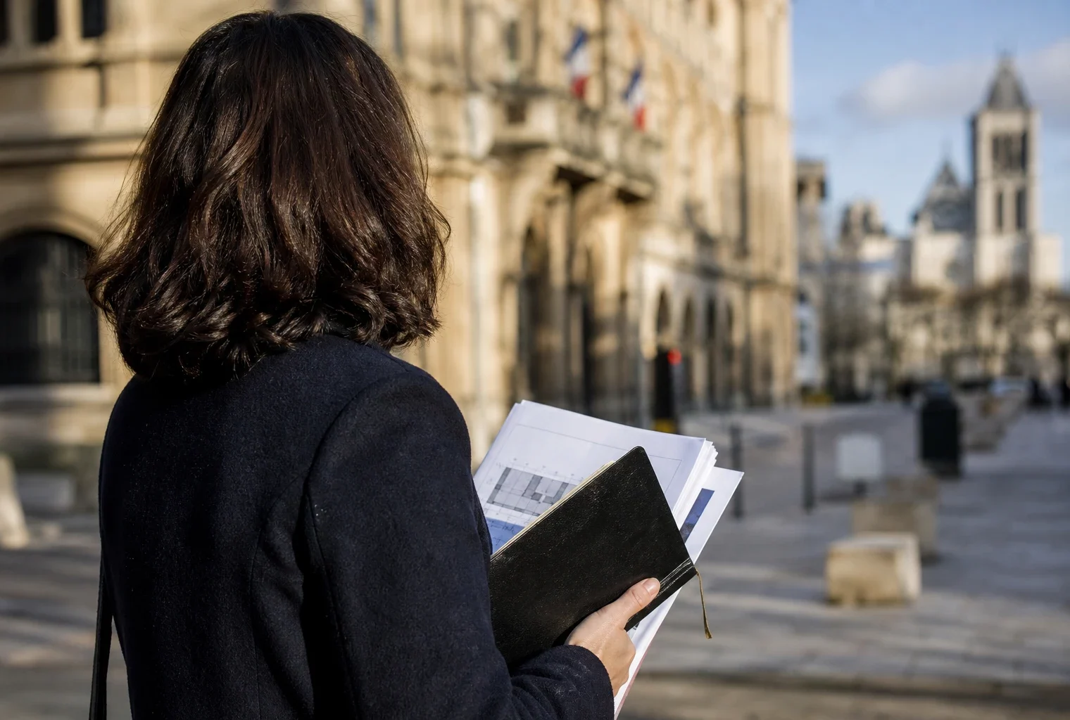 Un élu local anonyme prend des notes près de la mairie de Saint-Denis, avec la basilique floue en arrière-plan.