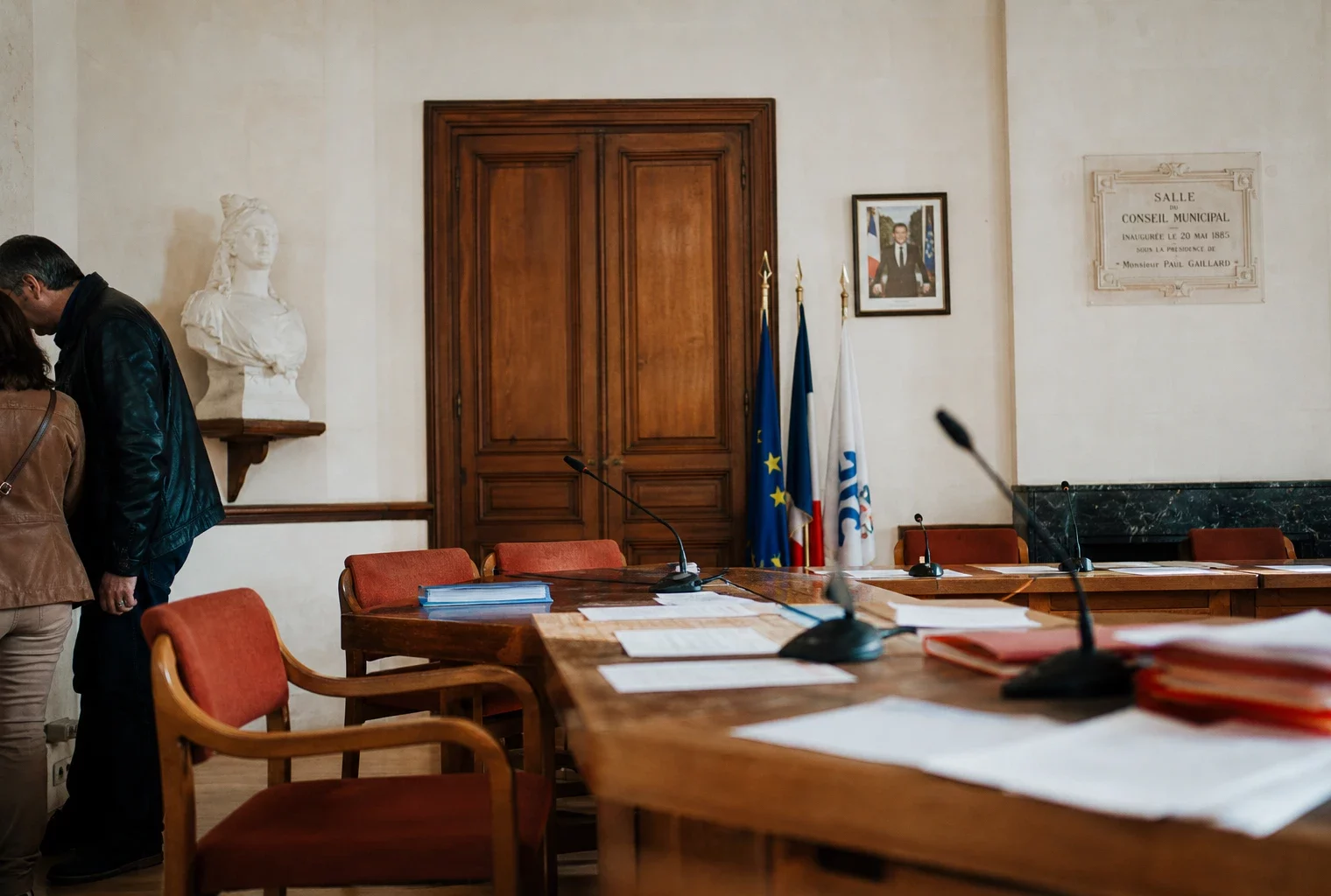 Salle de conseil municipal lumineuse avec chaise vide, micros et dossiers flous, en plan large oblique.