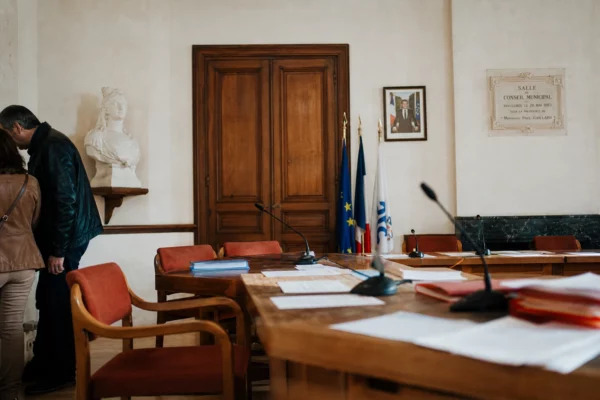 Salle de conseil municipal lumineuse avec chaise vide, micros et dossiers flous, en plan large oblique.