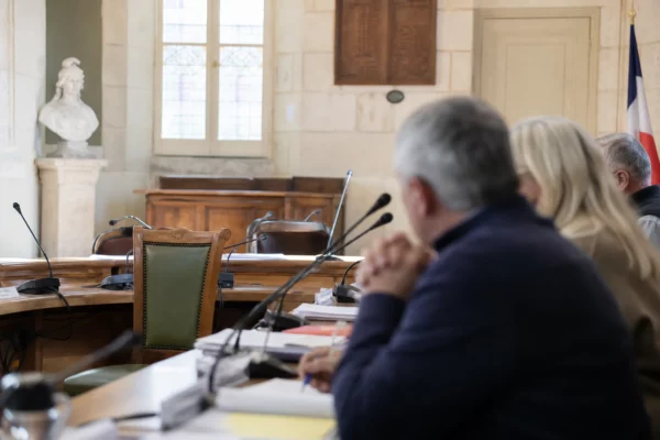 Salle de conseil municipal lumineuse avec chaise vide, micros et dossiers flous, ambiance de vote local.